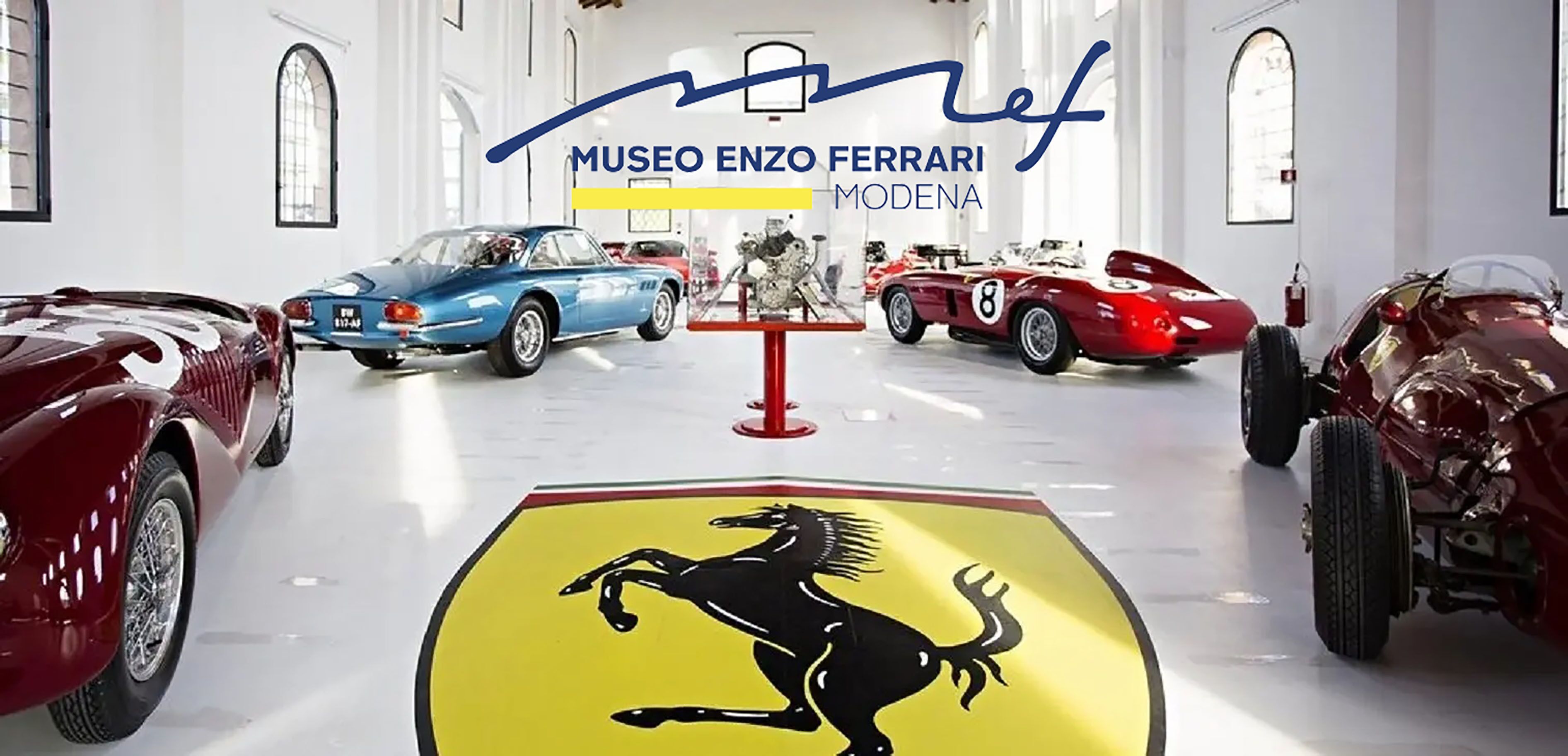 A museum interior with classic cars on display, a prominent Ferrari logo on the floor, and the text "MUSEO ENZO FERRARI MODENA".