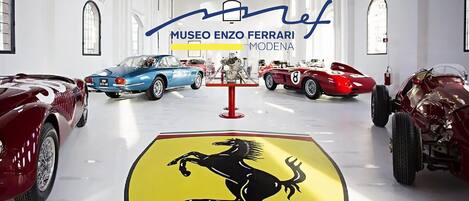En museiinteriör med klassiska bilar utställda, en framträdande Ferrari-logotyp på golvet och texten "MUSEO ENZO FERRARI MODENA".