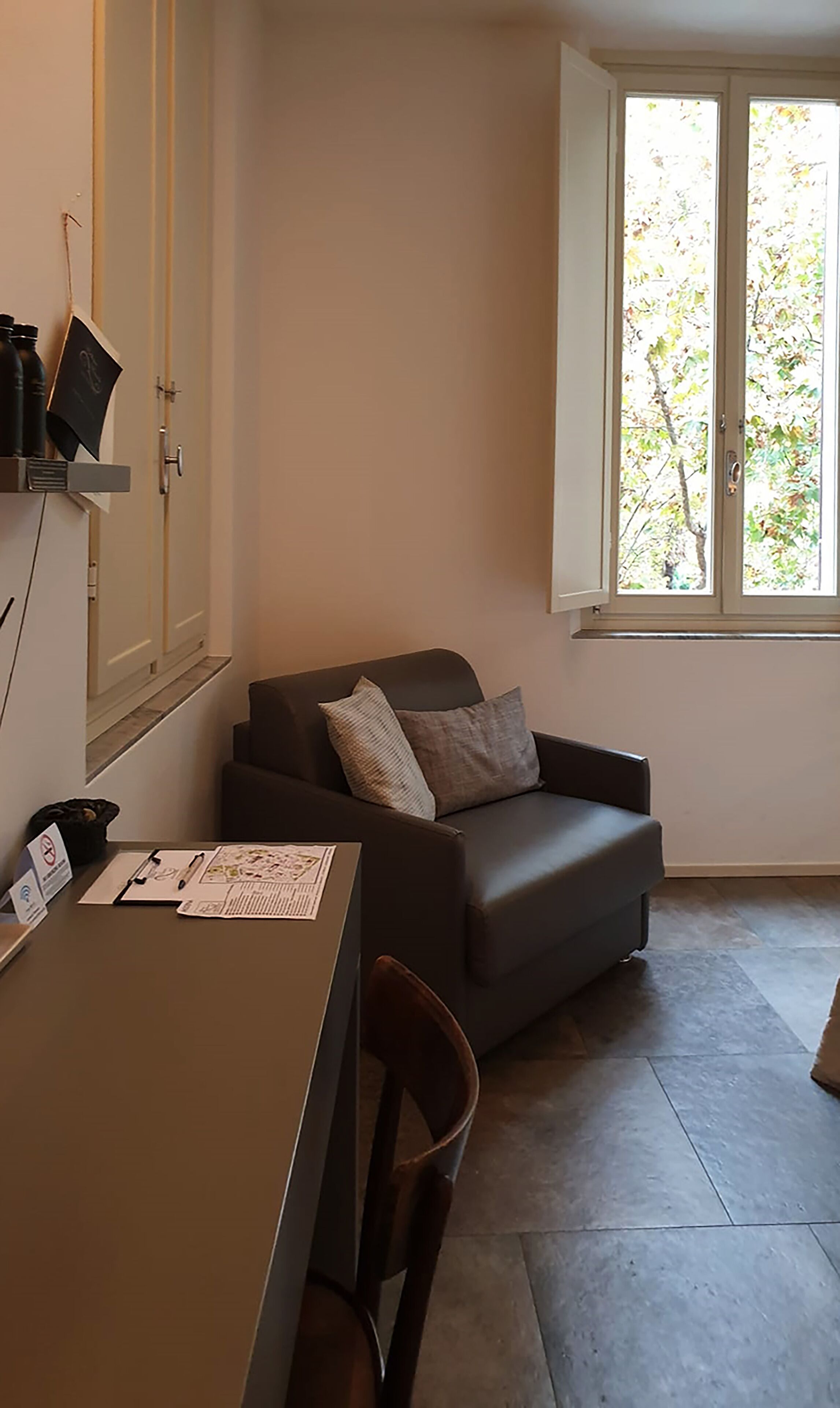 A small, modern room with a grey sofa, a wooden chair, a desk with papers, and a window with a view of trees.
