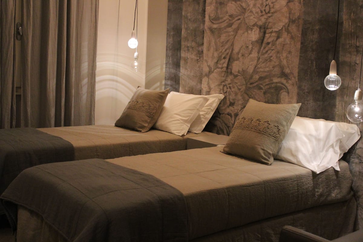 A hotel room with two beds, each with a brown bedspread and decorative pillows. There is a large window with curtains, and a wall with a textured pattern.