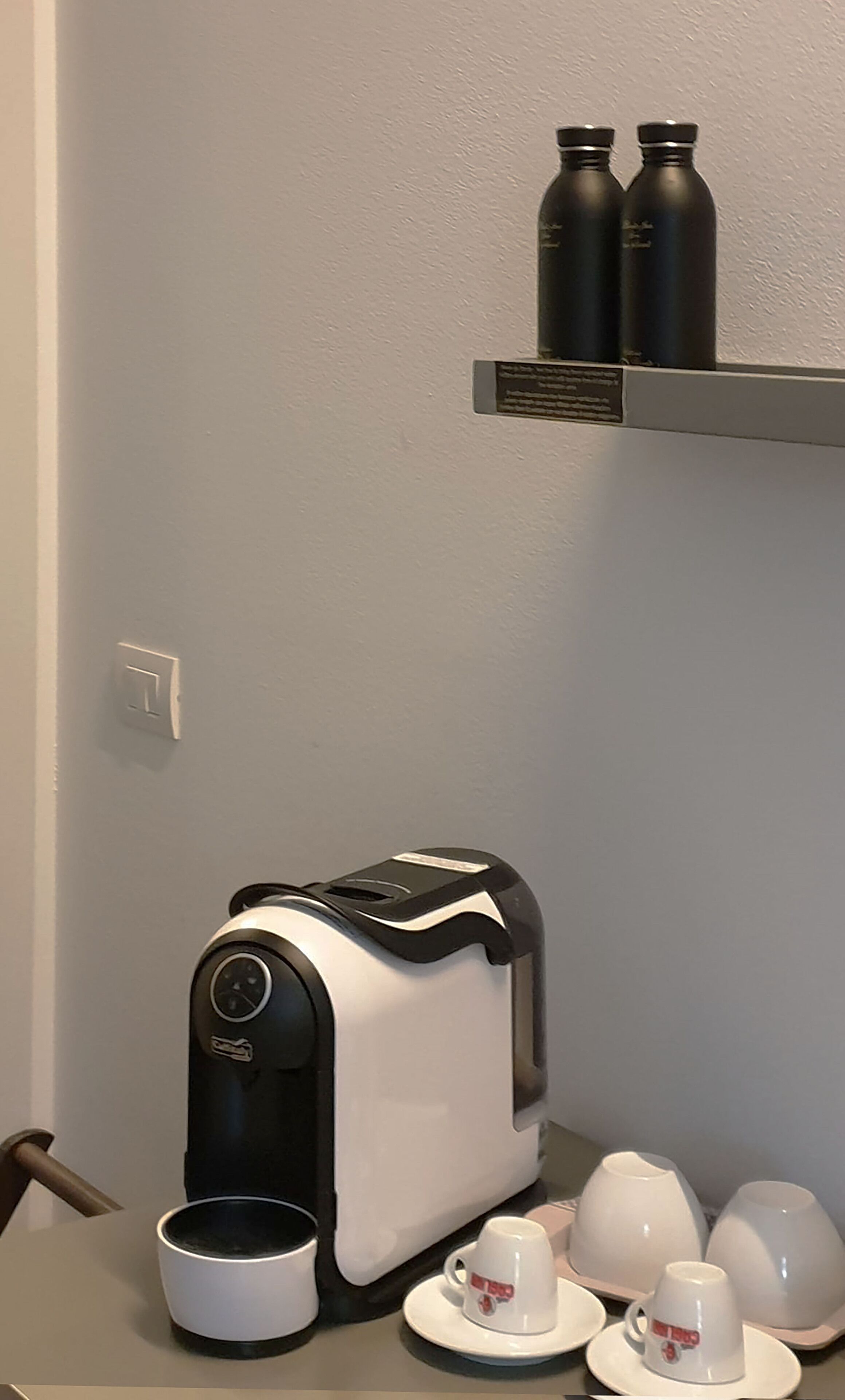 Una zona de cocina con una cafetera, dos termos y un juego de tazas y platillos blancos sobre una mesa.