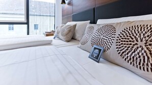 A modern bedroom with a large bed, decorative pillows, a bedside table, and a window with blinds. - elaya hotel munich city,  Trademark Collection by Wyndham (Munich)