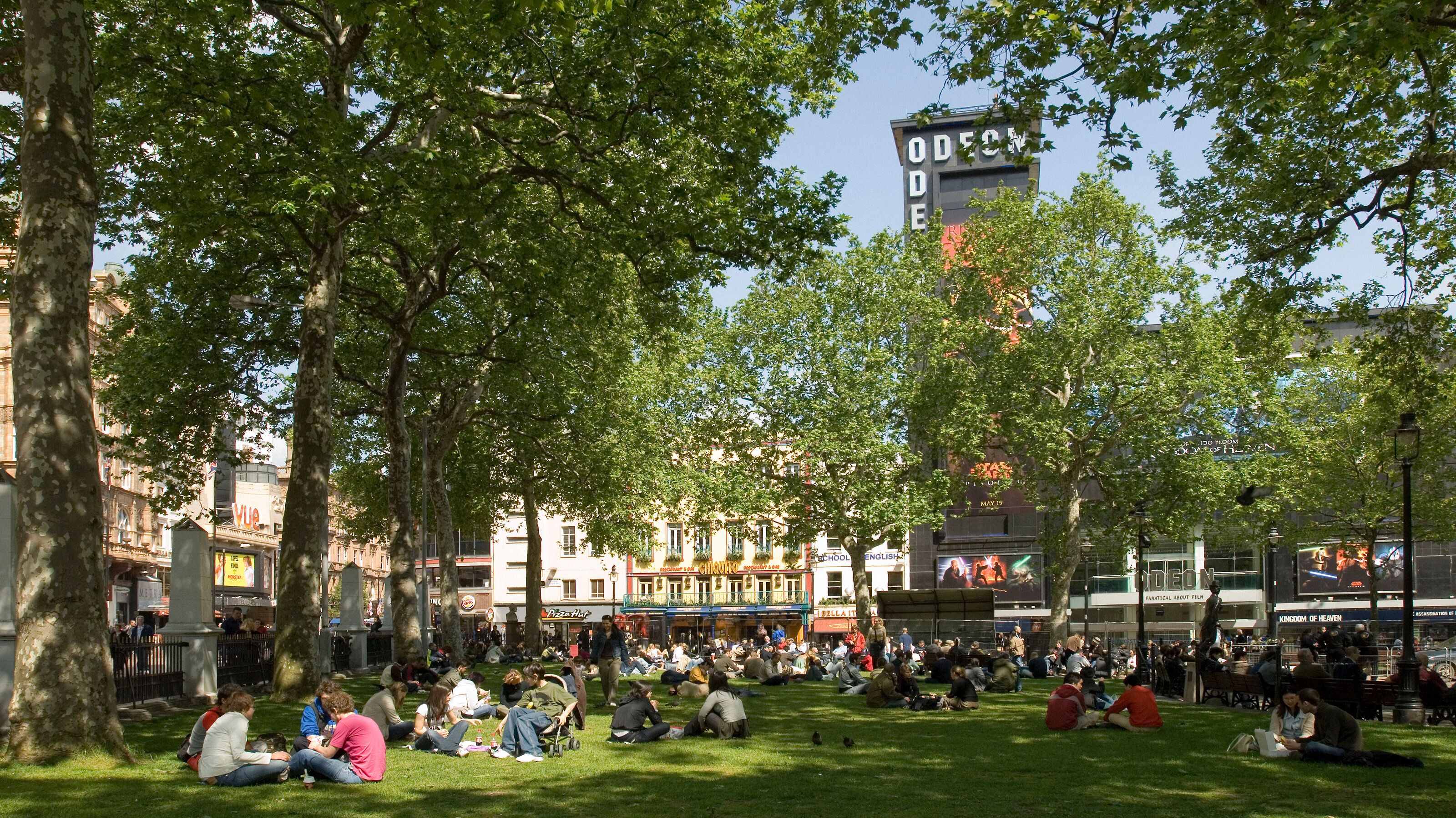 A park with people sitting on the grass, trees, and buildings in the background.
