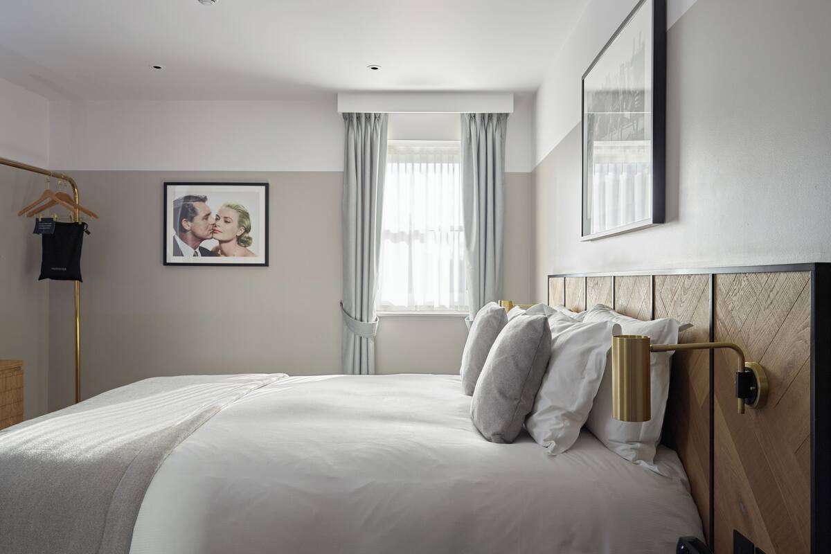 A hotel room with a bed, a bedside table with a phone, a picture on the wall, and a window with curtains.