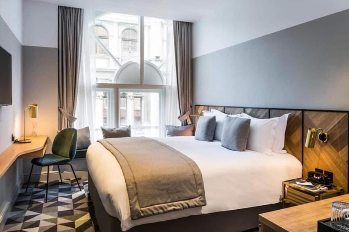 A modern hotel room with a large bed, a desk, a chair, and a view of the city through the window.
