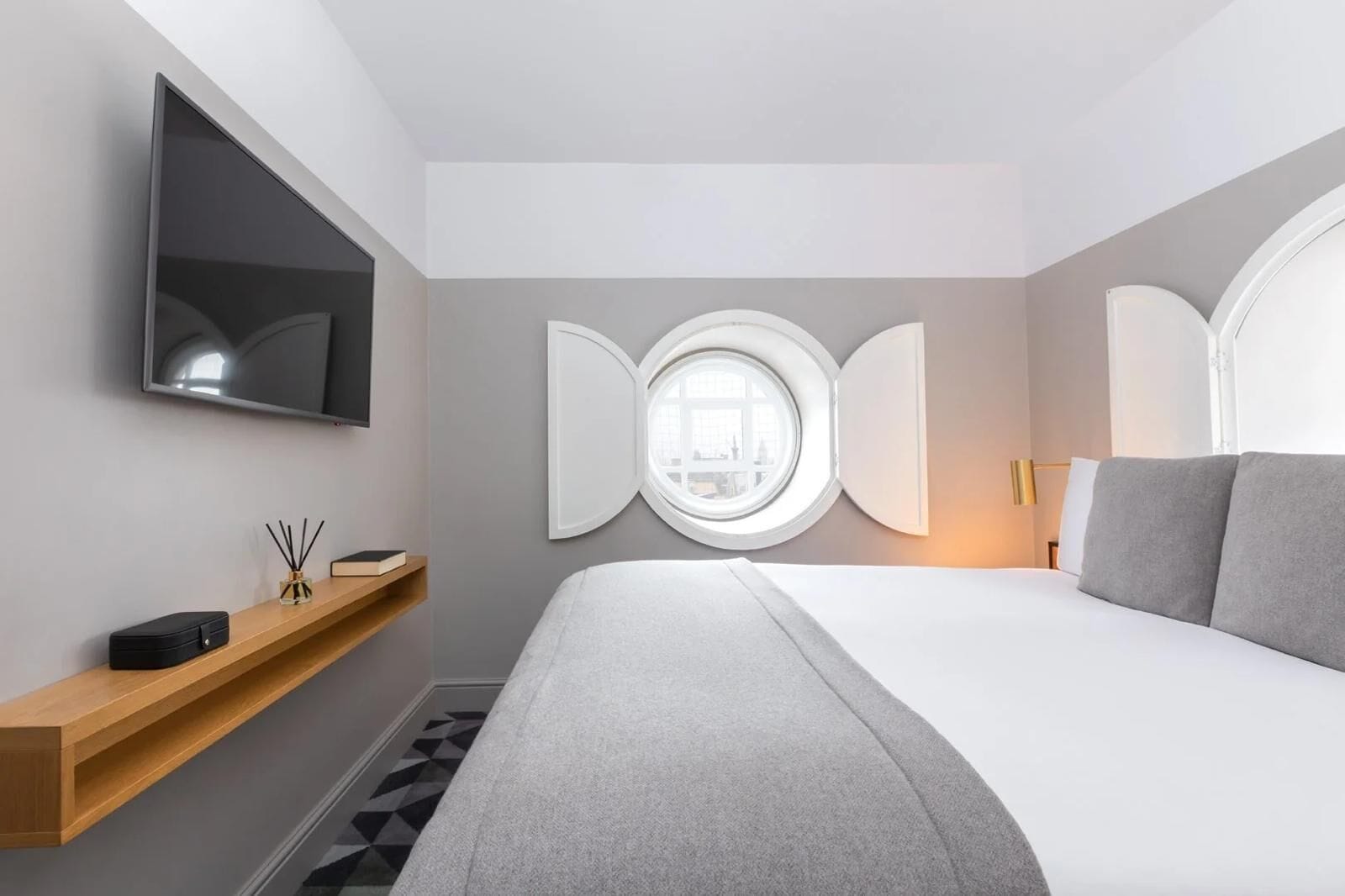 Een moderne hotelkamer met een groot bed, een bevestigde televisie, een houten plank met een boek en een zwarte tas, een rond raam en een wandlamp.