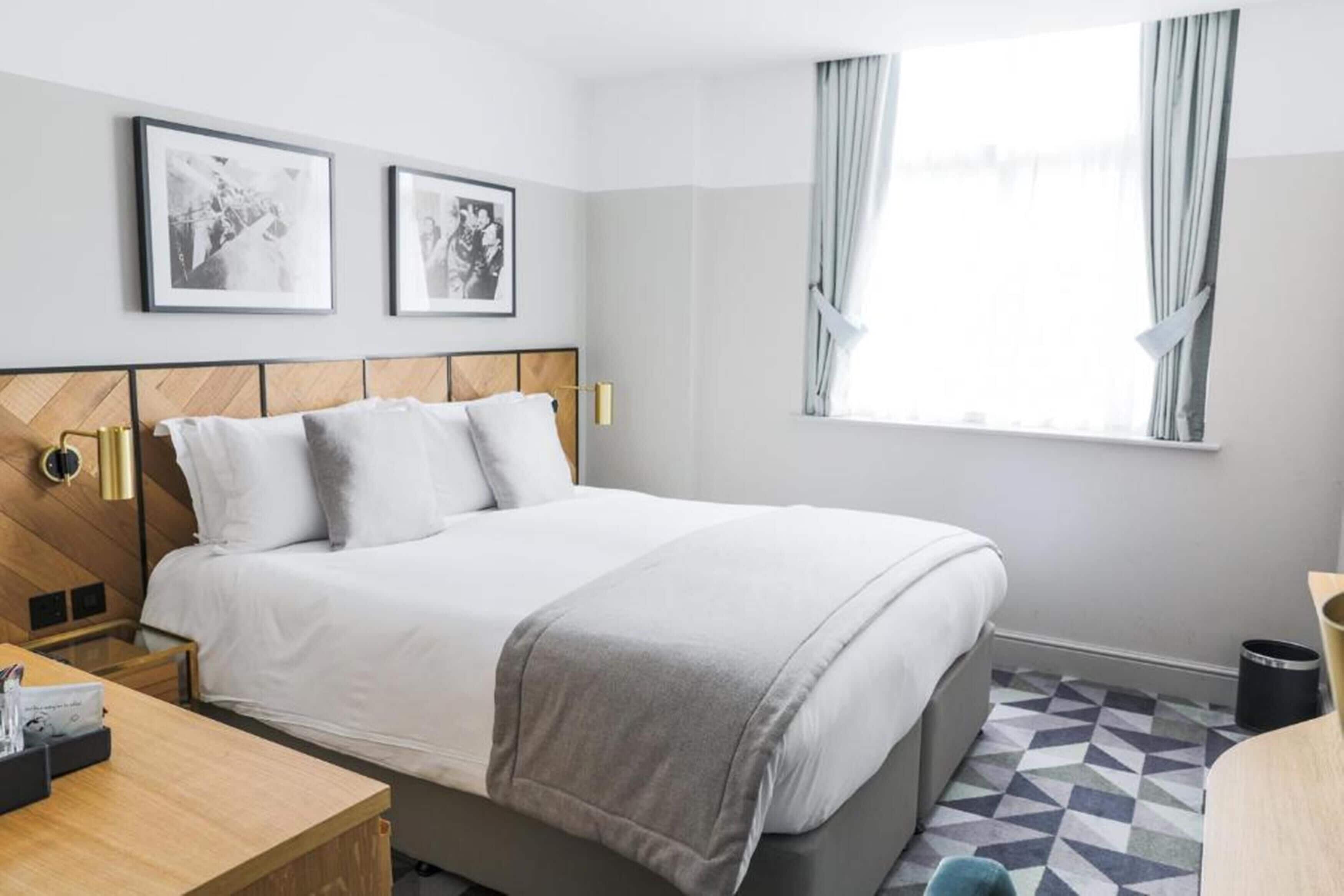 Een hotelkamer met een bed, nachtkastje, raam met gordijnen en twee ingelijste foto's aan de muur.