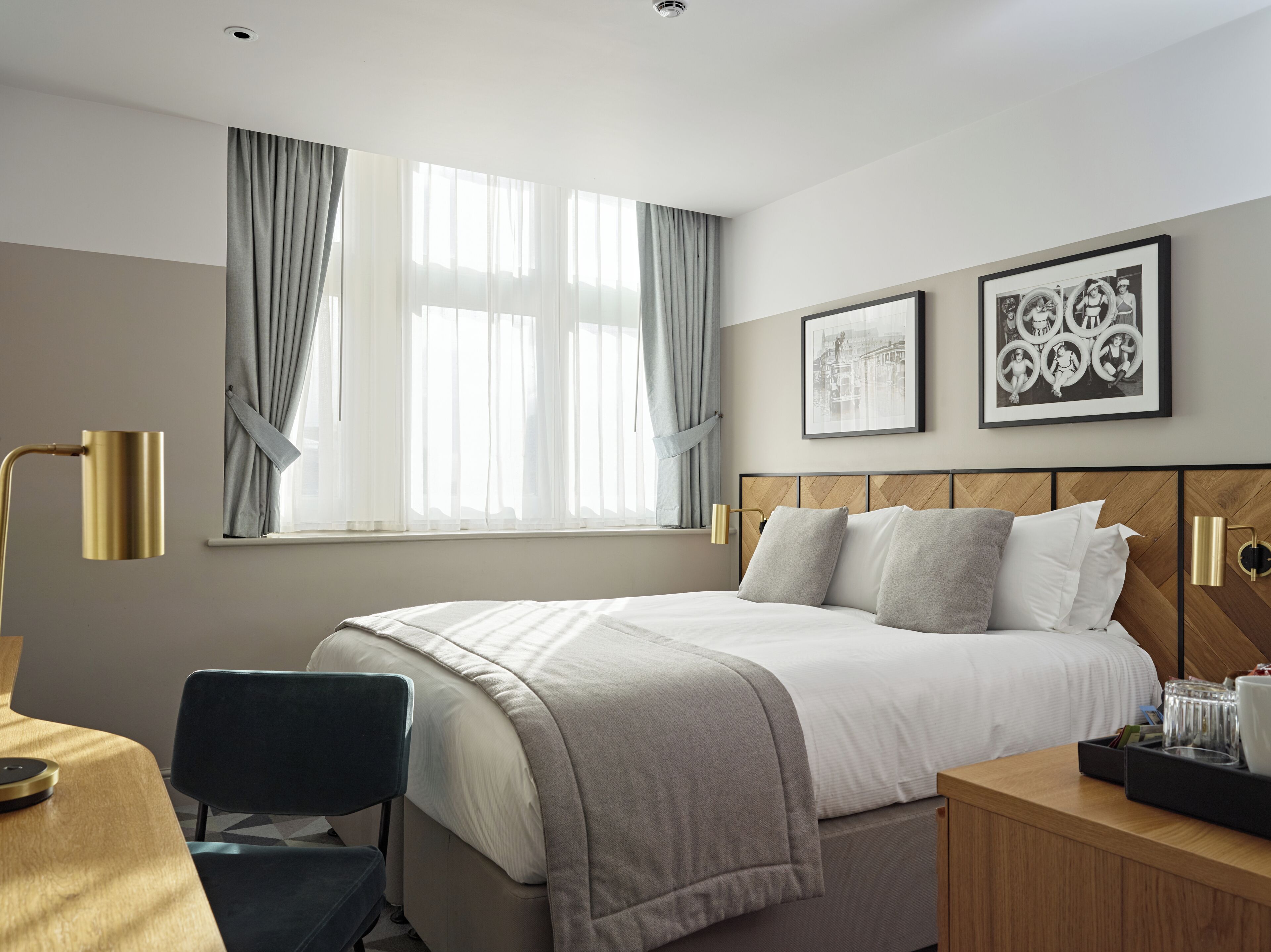 Ein Hotelzimmer mit einem Bett, einem Schreibtisch, einem Sessel, einem Fenster mit Vorhängen und zwei gerahmten Bildern an der Wand.