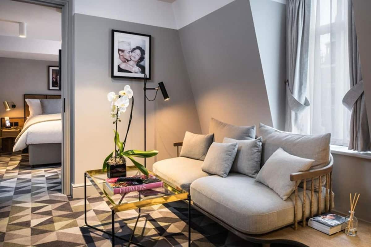 A modern hotel room with a sofa, a small table with a flower vase, a framed picture on the wall, and a bed in the background.