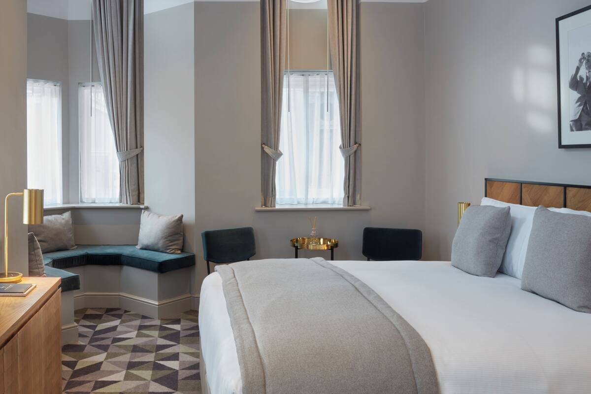 A modern hotel room with a large bed, a seating area with a view, and a desk with a lamp.