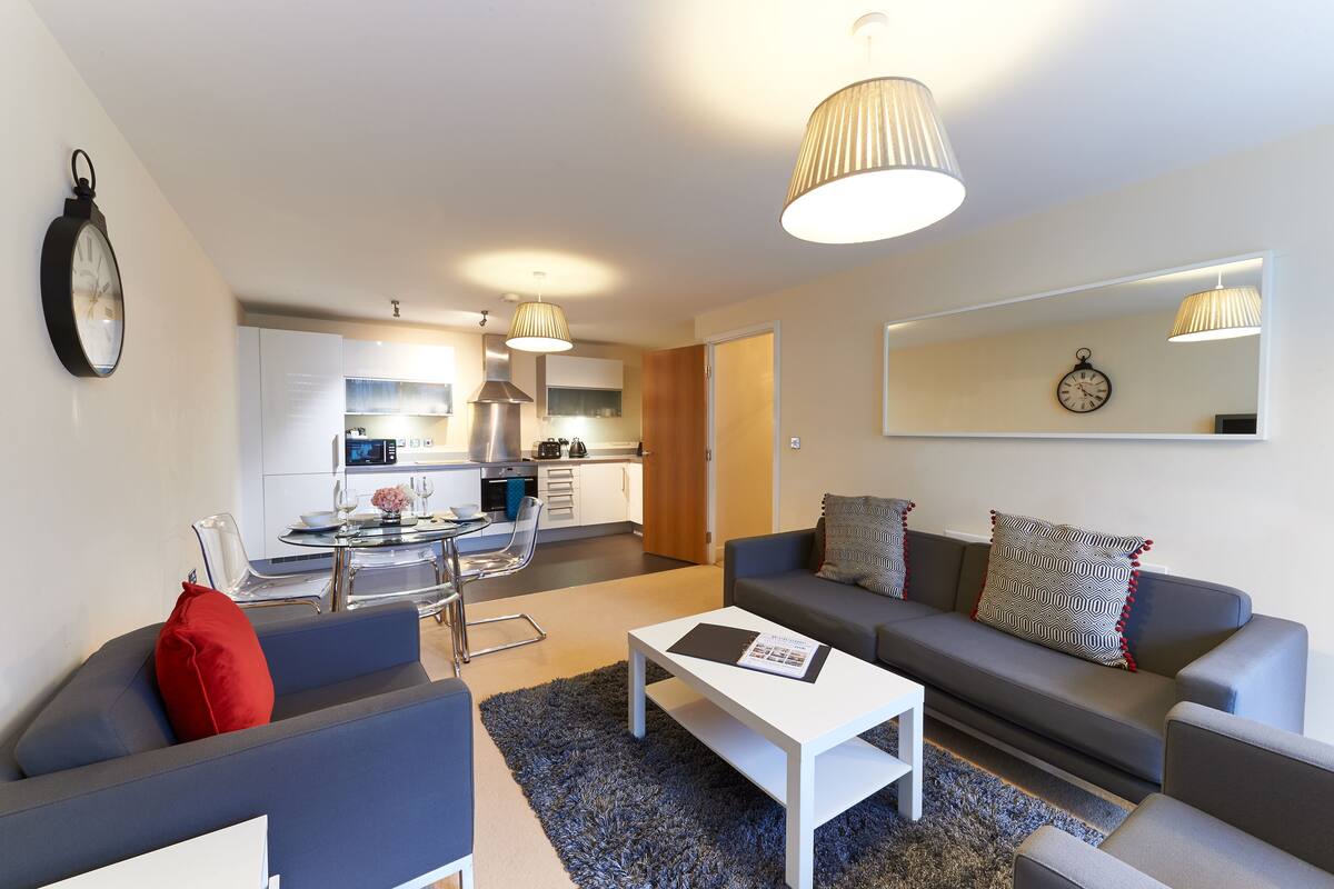 A modern living room with a grey sofa, a white coffee table, and a red pillow. There is a kitchen in the background with white cabinets and stainless steel appliances.