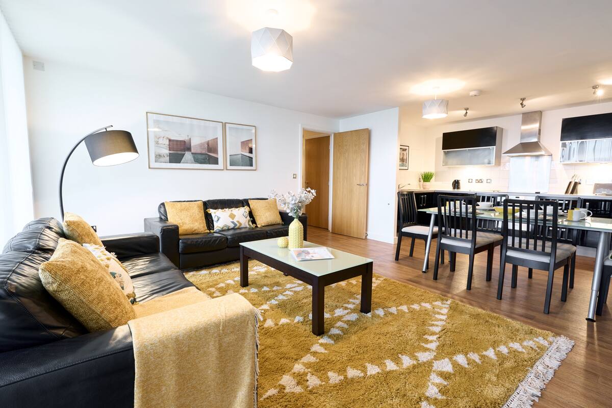 A modern living room with a black leather sofa, a coffee table, and a patterned rug. There is a dining area with a table and chairs, and a kitchen in the background.