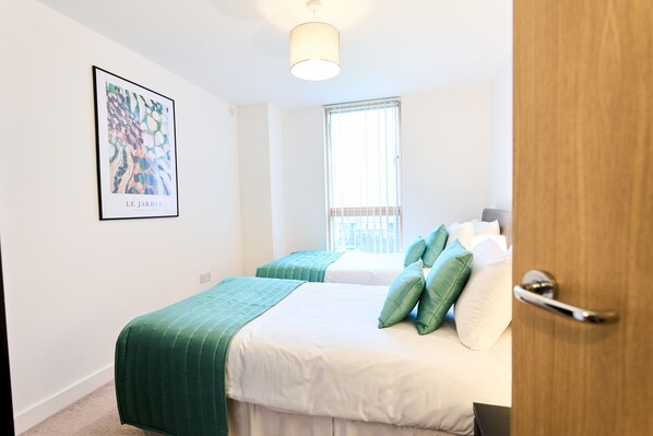 A bedroom with two beds, a window with blinds, a painting on the wall, and a door. - Vizion by ShortstayMK (Milton Keynes)