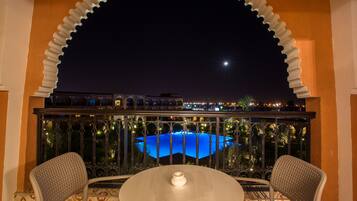 A balcony with a view of a lit swimming pool at night, surrounded by ornate railings and two chairs.