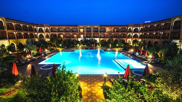 A large outdoor swimming pool surrounded by lounge chairs and umbrellas, with a building featuring arched balconies in the background. - Ennakhil Marrakech Hotel & SPA (Marrakech)