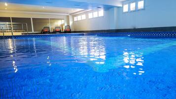An indoor swimming pool with clear blue water, a diving board, and three sun loungers with orange cushions.
