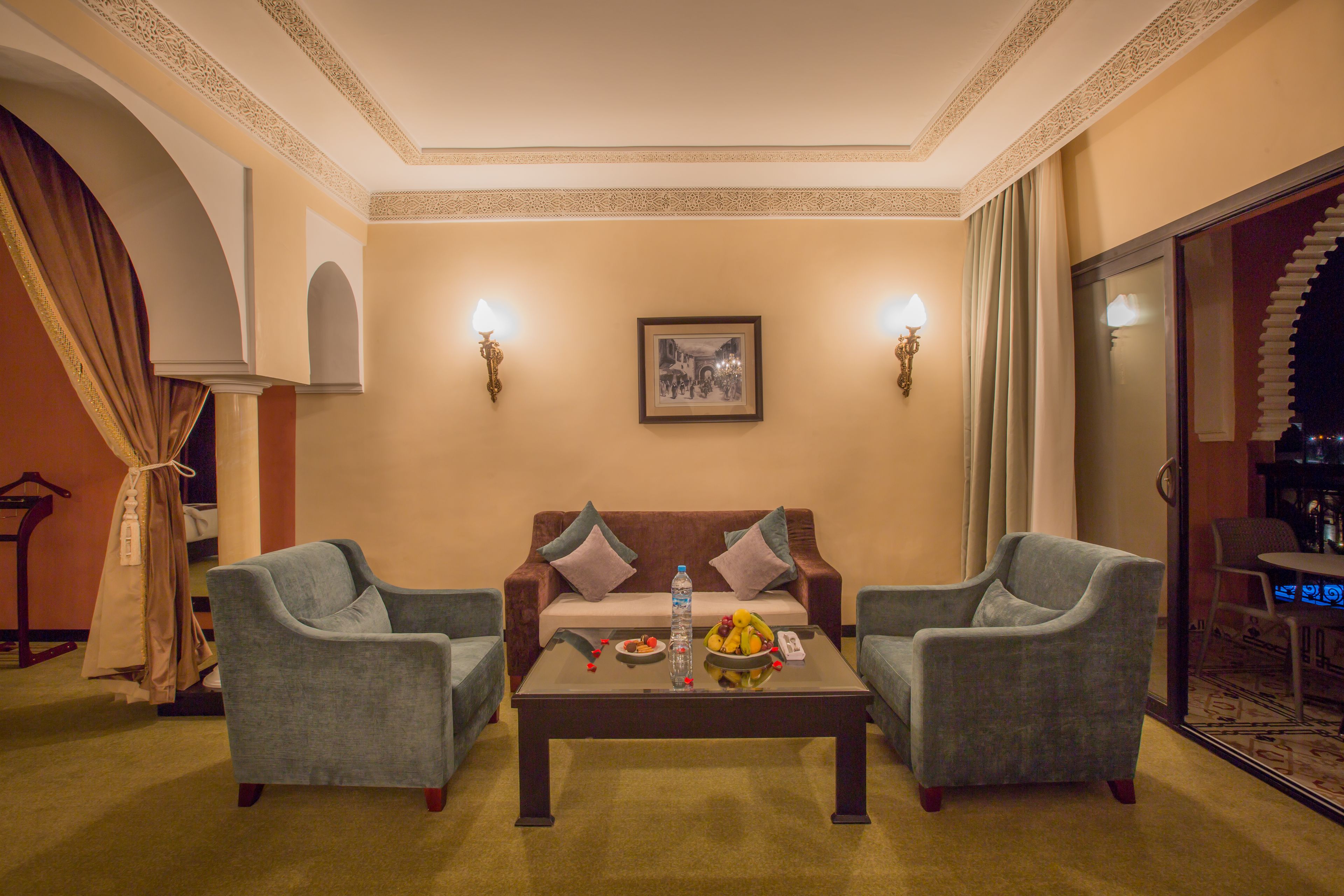 A living room with a sofa, two armchairs, a coffee table with fruits and a bottle, and a painting on the wall.