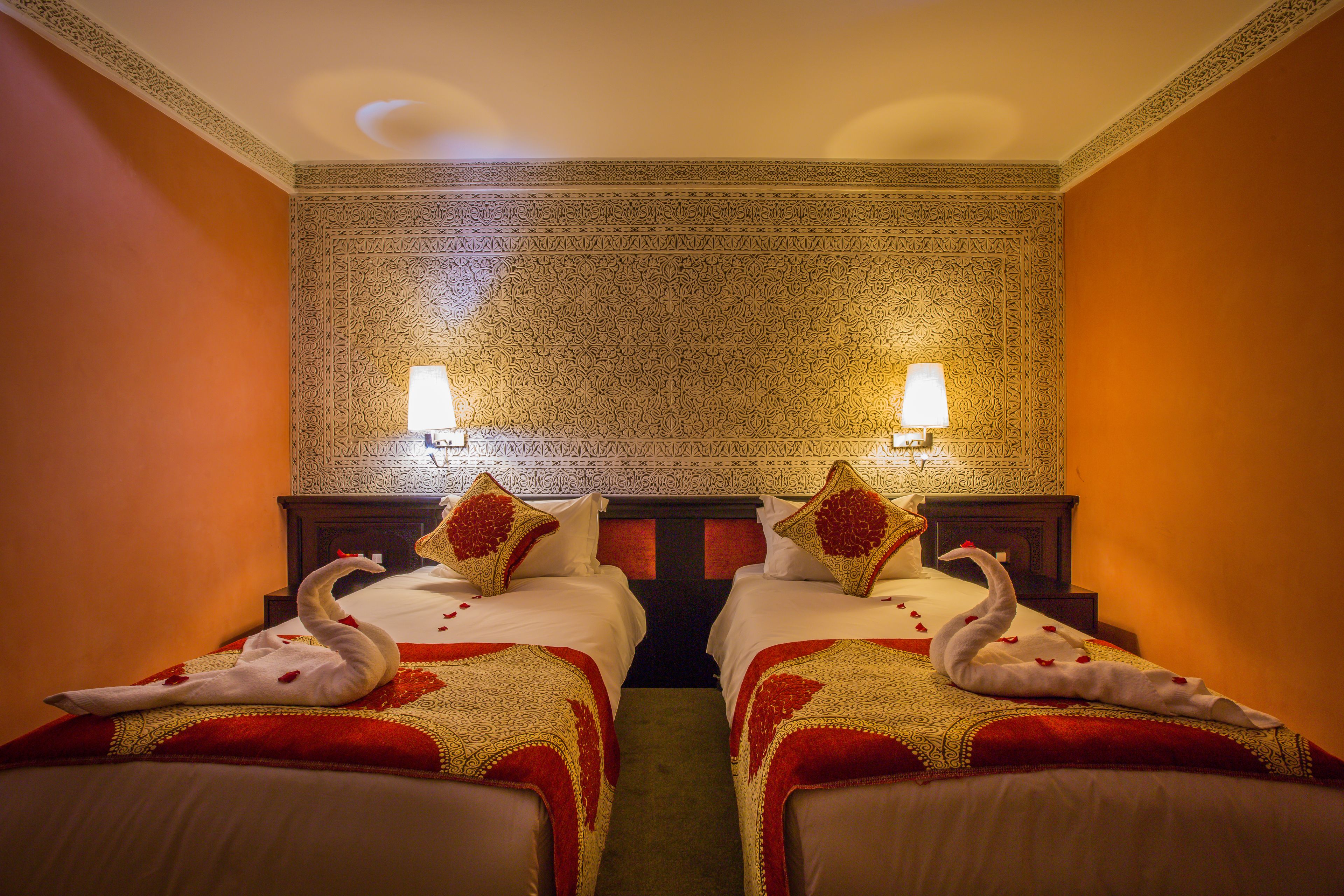 Camera d'albergo con due letti, ognuno decorato con una coperta a forma di cigno e petali di rosa. La stanza presenta una carta da parati decorata e due lampade fissate alla parete.