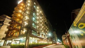 A multi-story building with illuminated windows and balconies, a parking area, and a street with moving vehicles. - Wsotel Hotel and Serviced Apartment (Songkhla)