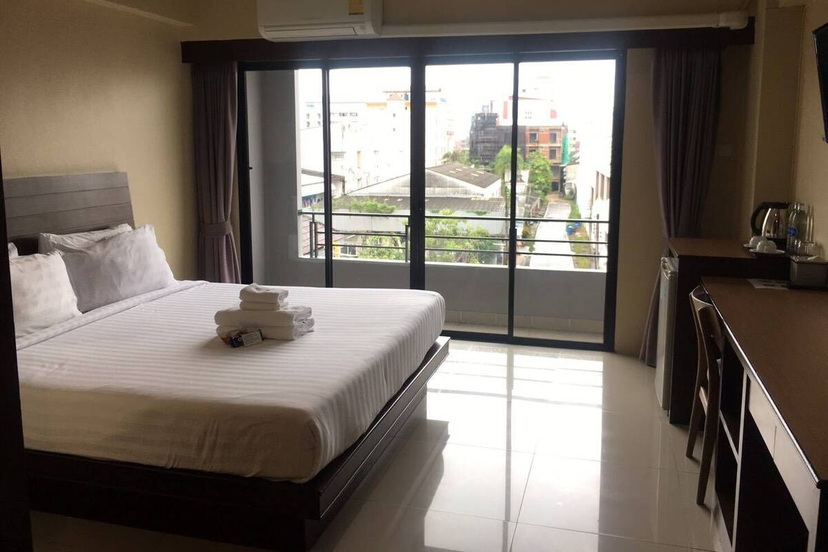 Une chambre d’hôtel équipée d’un lit, d’un bureau, d’une chaise, d’un appareil de climatisation et offrant une vue sur les bâtiments par le biais d’une grande fenêtre.