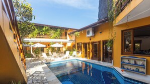 Une piscine entourée de chaises longues et de parasols, elle-même bordée d’une terrasse dallée de pierres et d’un bâtiment jaune.
