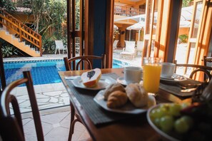 Ein Frühstückstisch mit Blick auf einen Pool, gedeckt mit Croissants, Obst und Saft, sowie ein Stuhl.