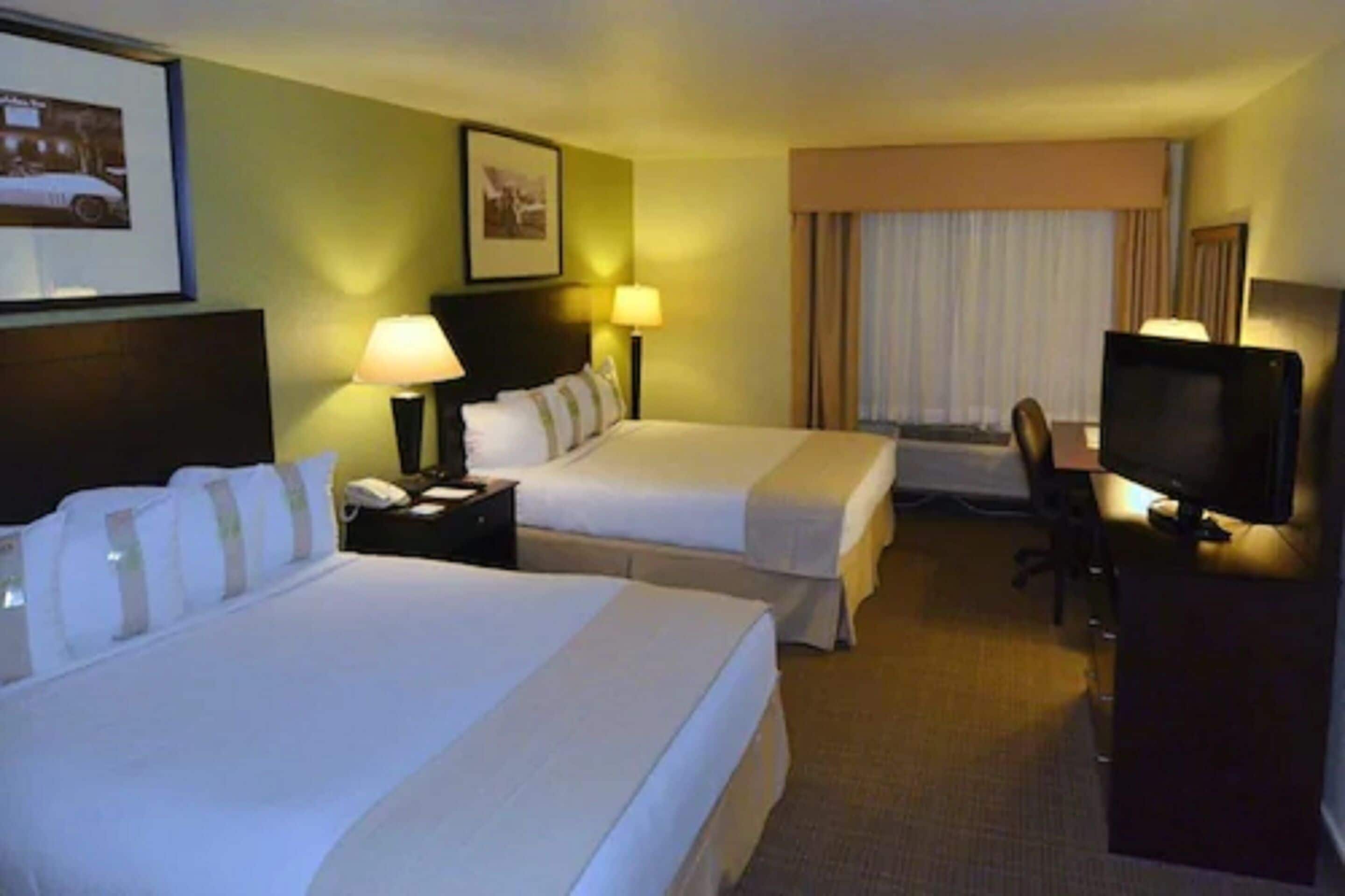 A hotel room with two beds, a desk with a computer, a TV, and a window with curtains.