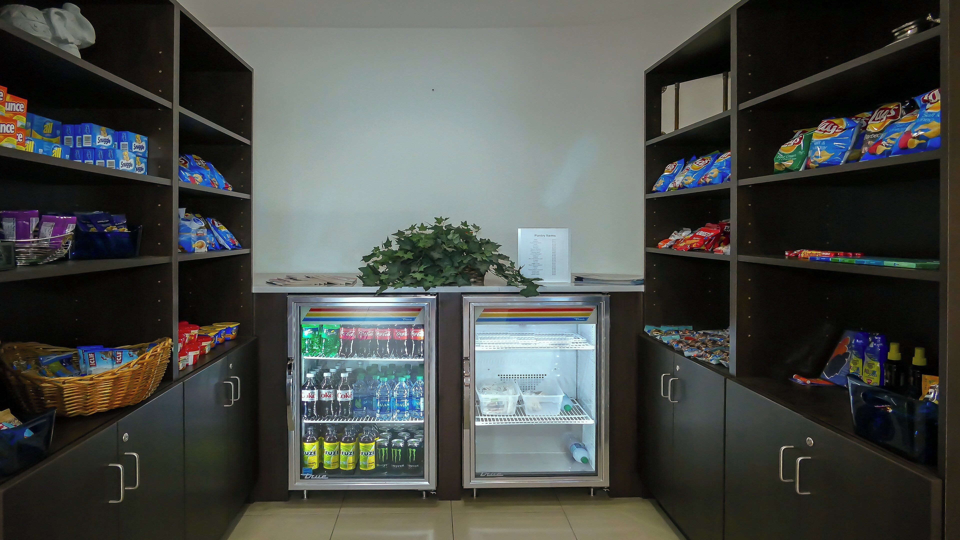 A small kitchenette area with two beverage dispensers, shelves stocked with snacks, and a basket of items on the counter.