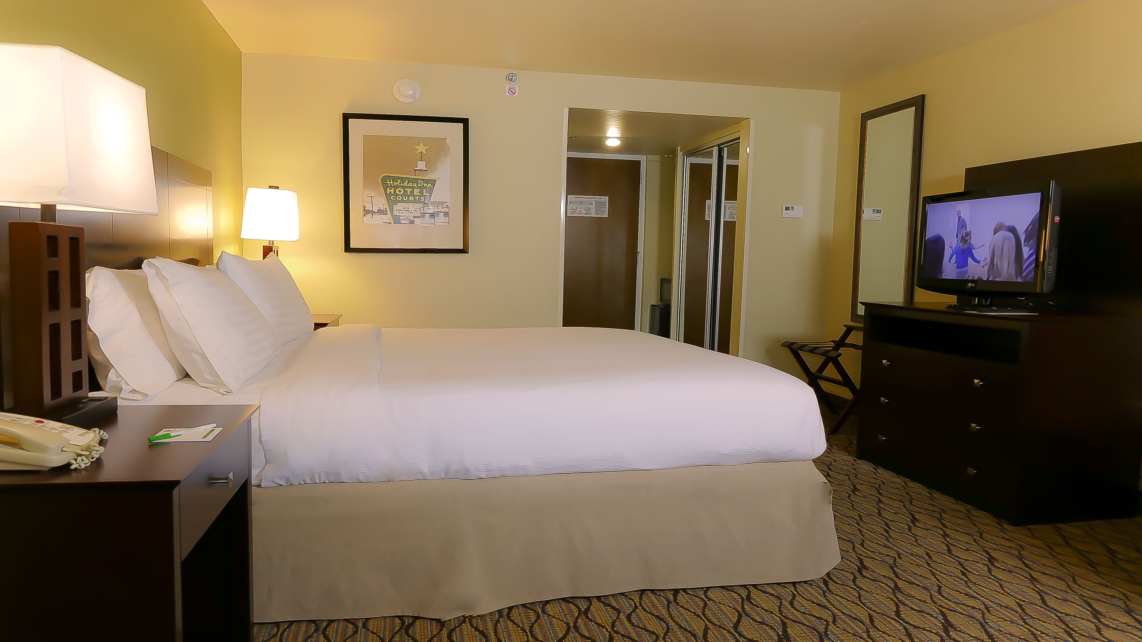 Quarto de hotel com uma cama, uma escrivaninha com telefone, televisão e um quadro na parede.
