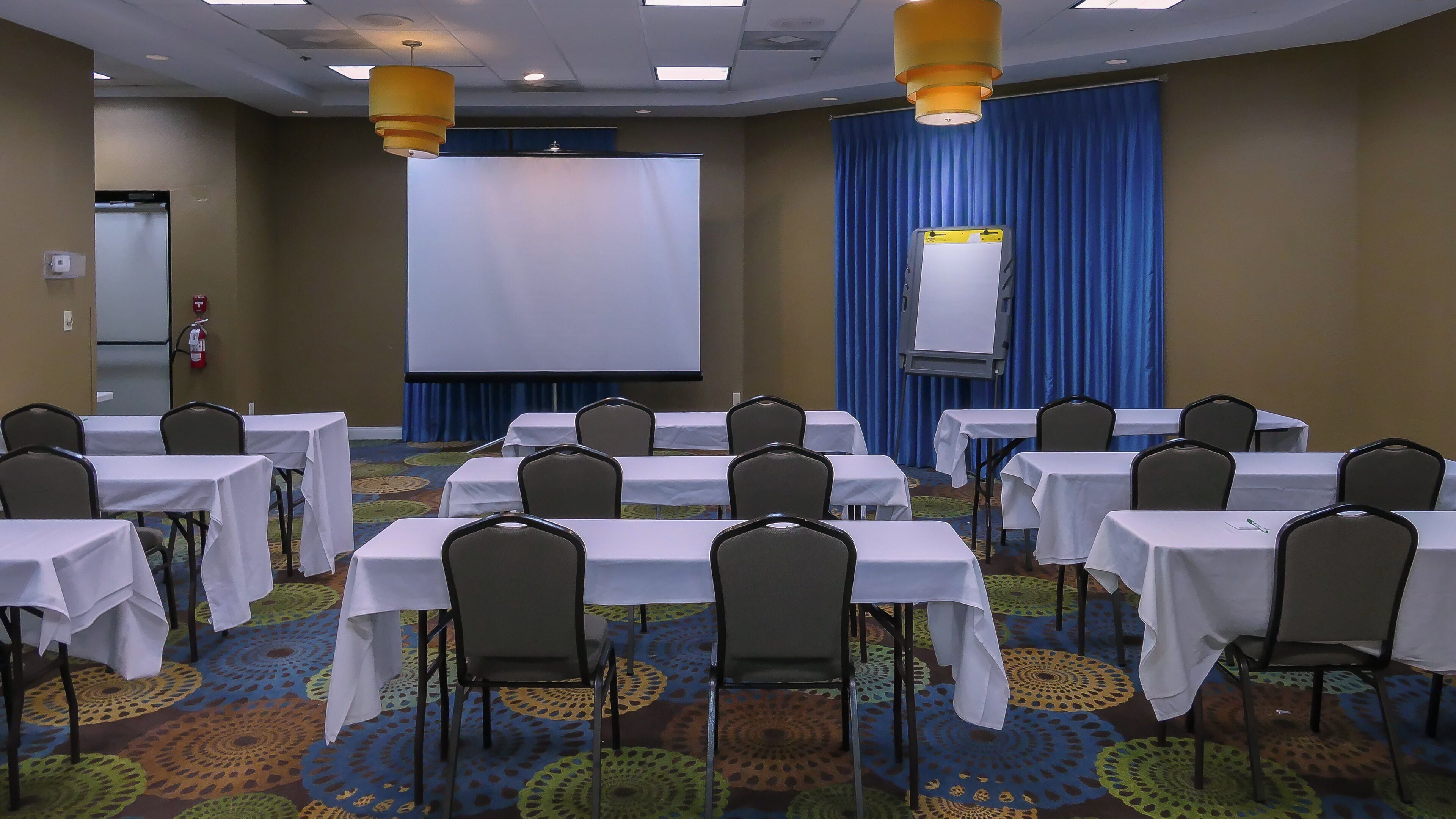 Un salón de conferencias con mesas, sillas, una pantalla de proyector y un telón de fondo azul.