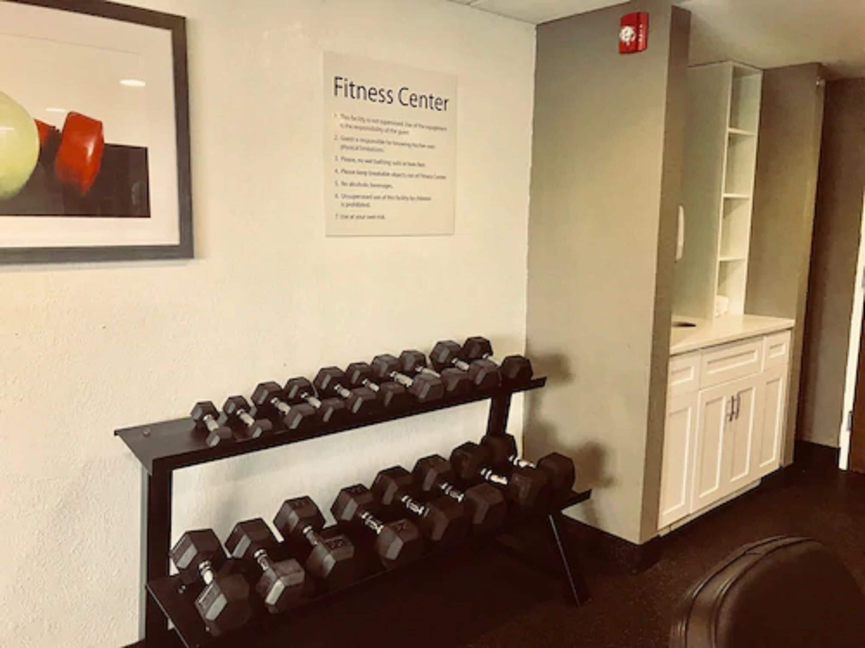 Un gimnasio con un estante de mancuernas y un cartel con las normas.