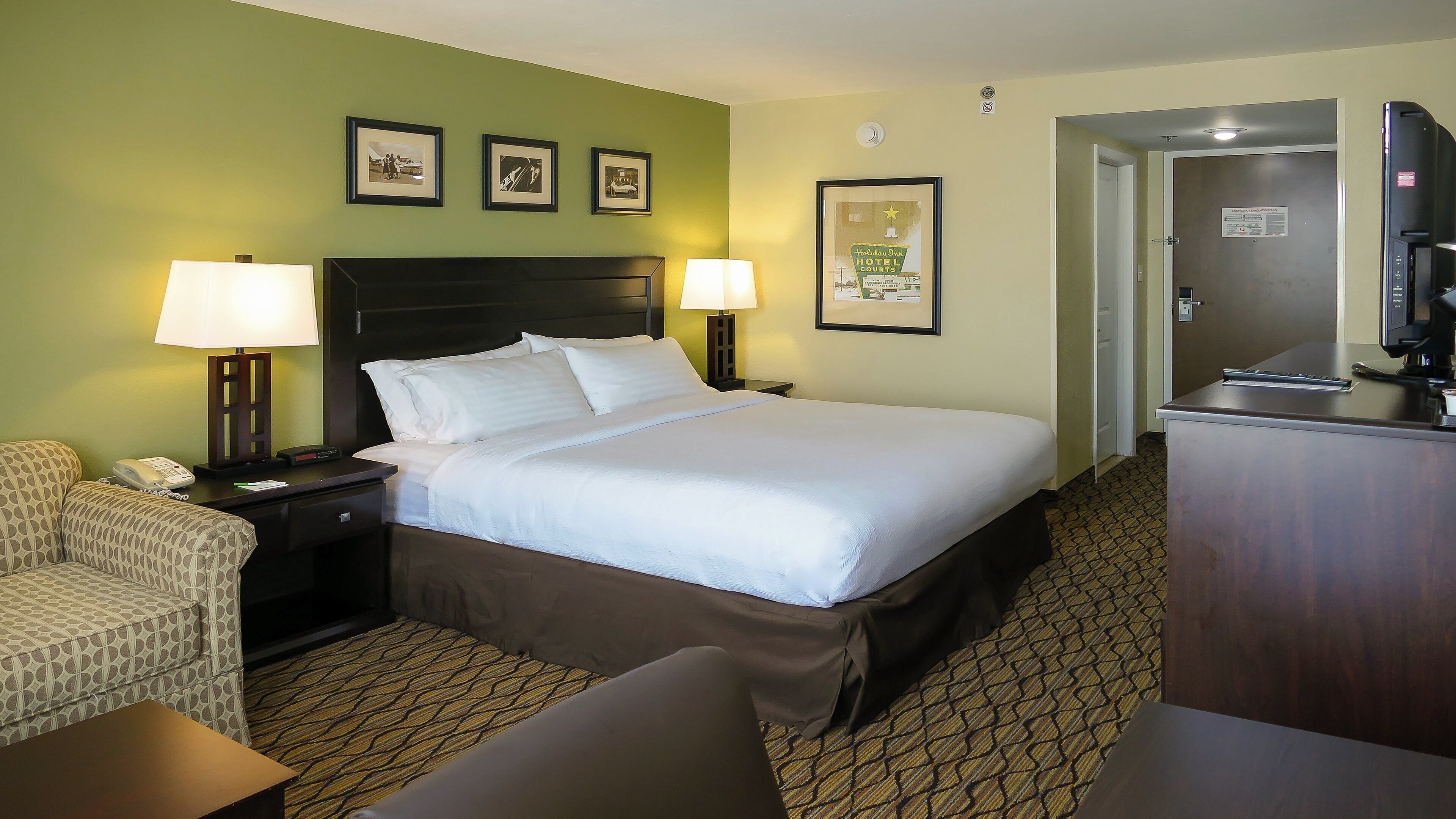 Quarto de hotel com uma cama, duas luminárias de cabeceira, uma escrivaninha com telefone, uma cadeira e uma televisão.