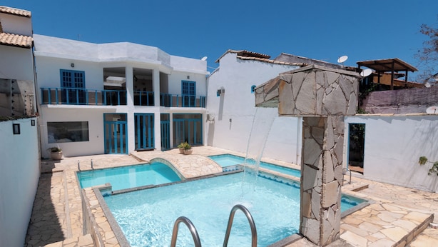 Una piscina con baldosas de piedra, un pilar de piedra y un edificio blanco con puertas y ventanas azules.