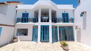 Un edificio blanco de dos pisos con ventanas y puertas con marcos azules, un balcón y un patio adoquinado.