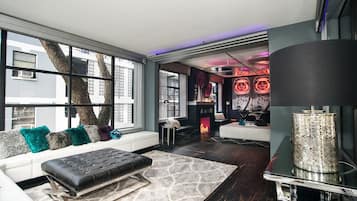 A modern living room with a large window, a fireplace, and a seating area with a black ottoman.