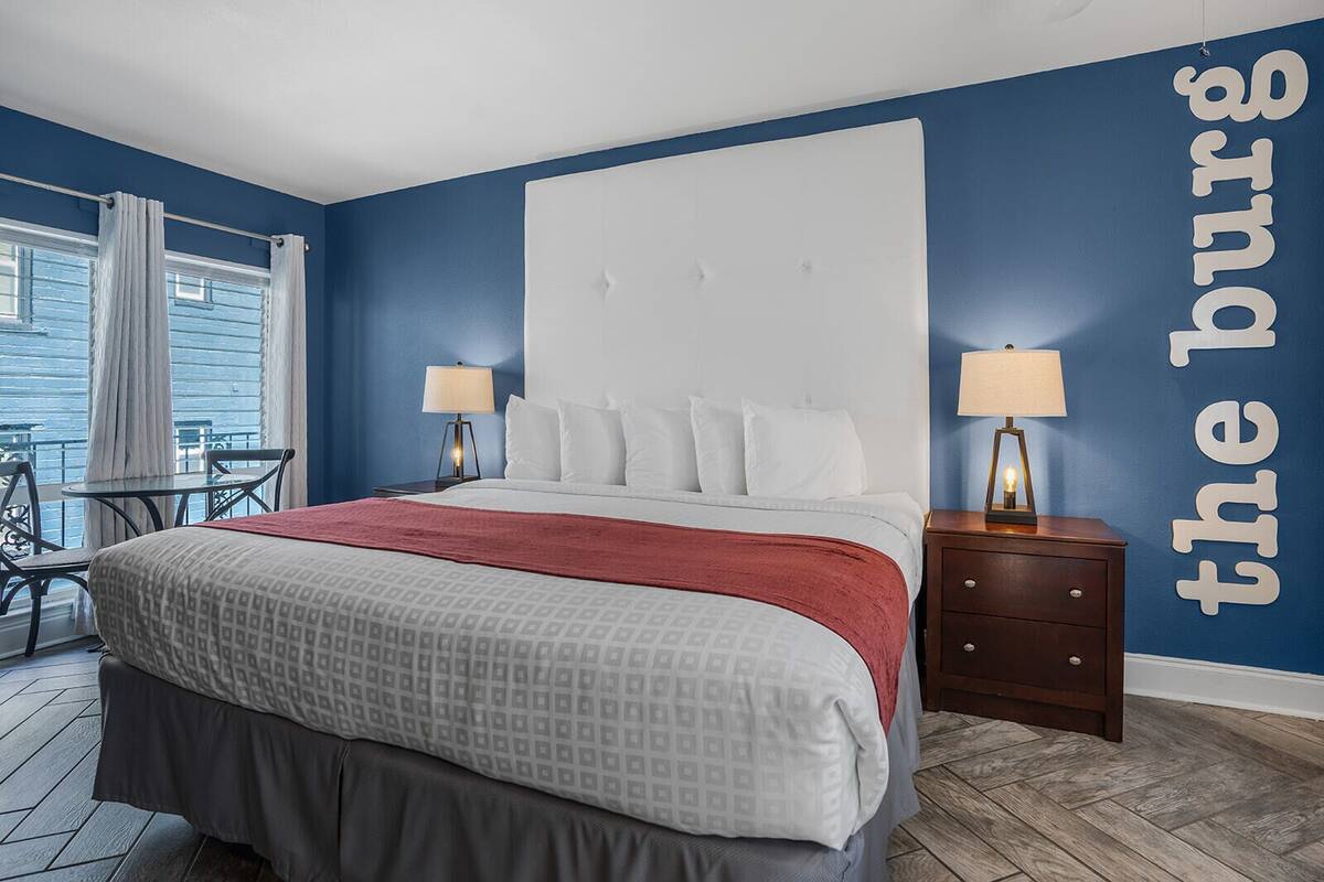 A bedroom with a bed, two bedside tables with lamps, a window with curtains, and a view of the water outside.
