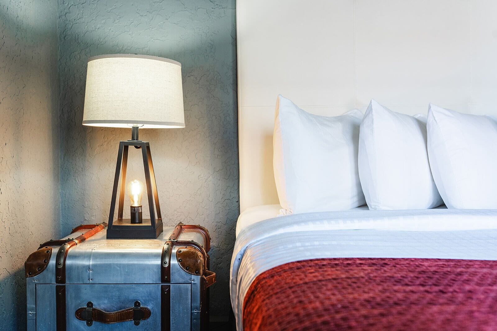 A bed with white pillows and a red blanket, a vintage-style lamp on a metal suitcase, and a textured wall.