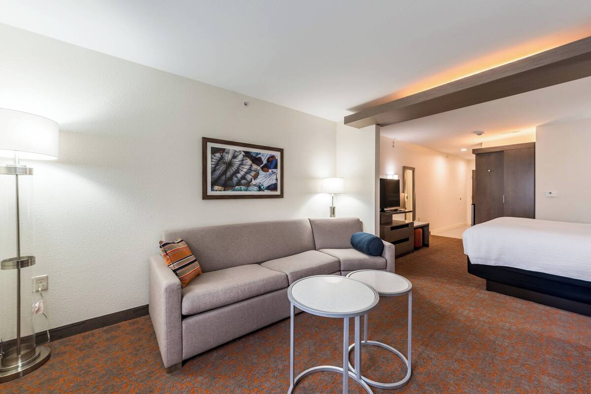 Habitación de hotel con sofá, dos mesas redondas, cama, lámpara y un cuadro en la pared.