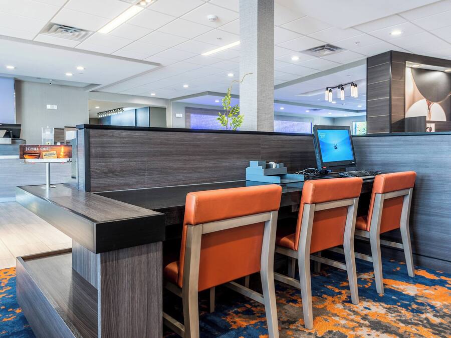 Un moderno vestíbulo de hotel con mostrador de recepción, una zona de estar con sillones naranja, una estación de computadoras y una planta decorativa.
