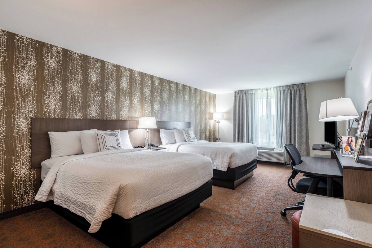 Habitación de hotel con dos camas, un escritorio, una silla y un ventanal con cortinas.