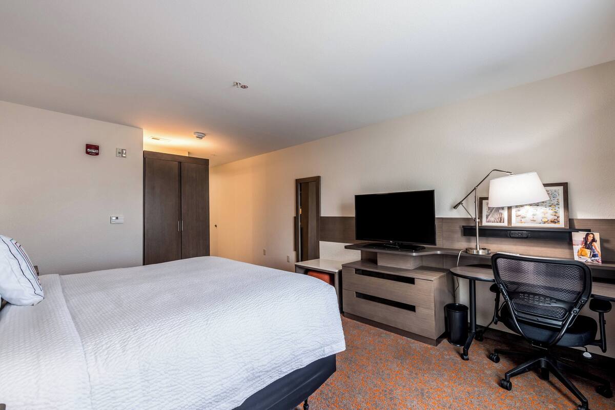 Habitación de hotel con cama, escritorio, silla, televisión y una kitchenette pequeña.