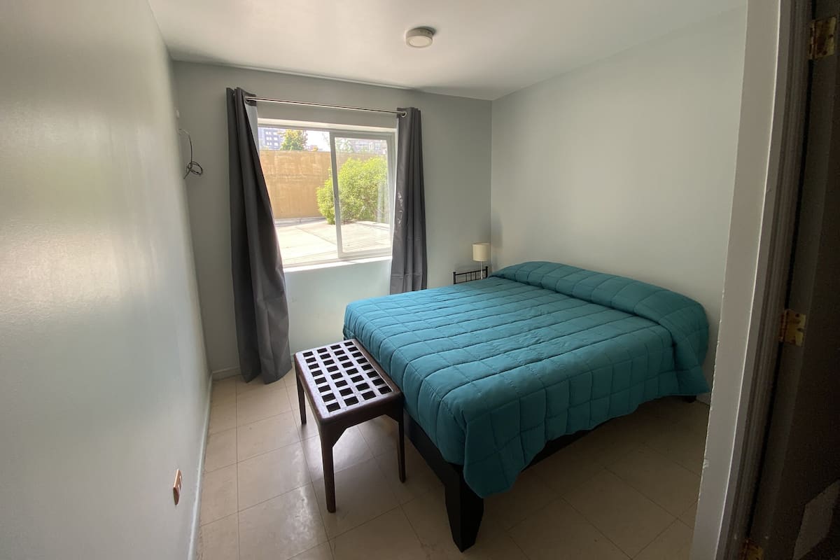 Un dormitorio con cama, una mesita, una ventana con cortinas y una lámpara.