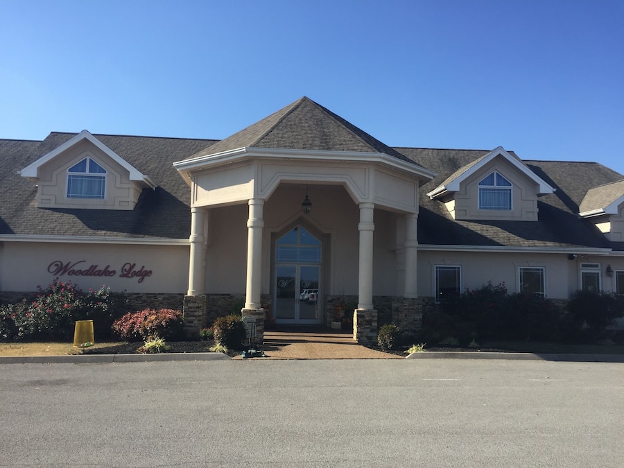 A two-story building with a prominent entrance under a covered porch, a sign reading "Woodlake Lodge," and a clear blue sky.