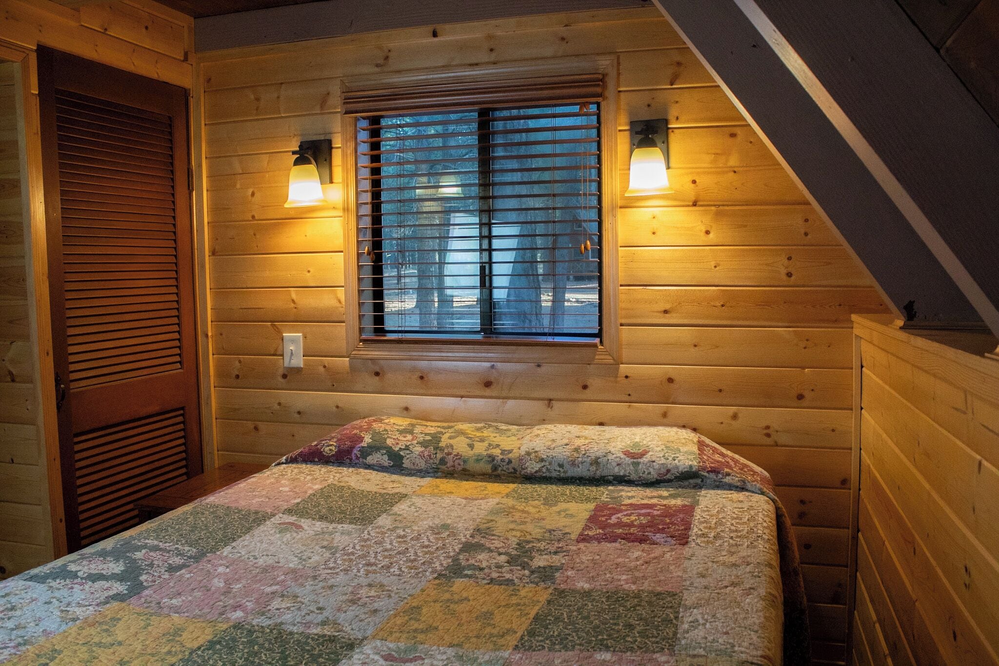 A wooden cabin bedroom with a bed, a window with blinds, and two wall-mounted lamps.