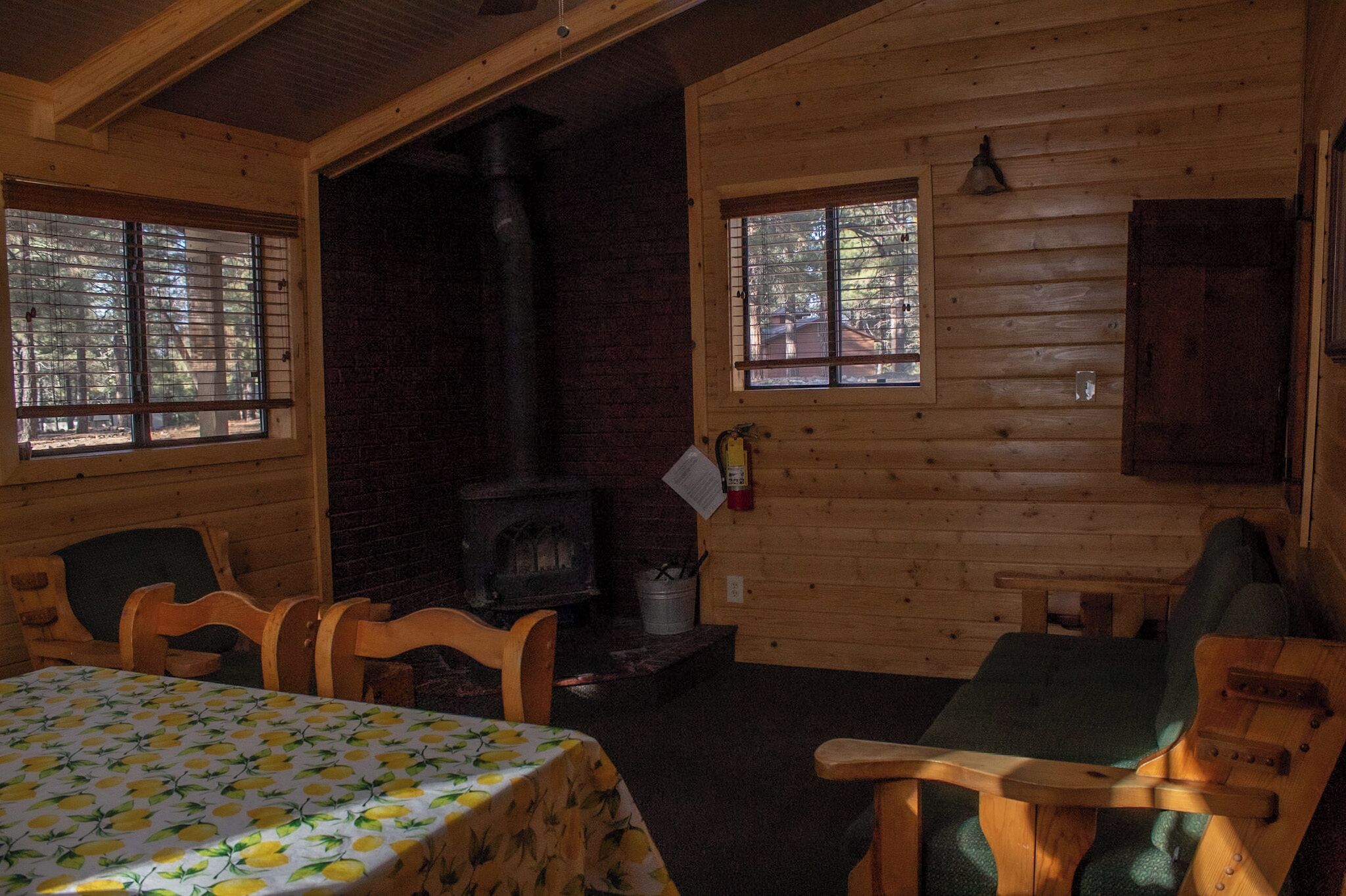 A cozy cabin interior with a fireplace, wooden walls, a table with a lemon pattern tablecloth, and two chairs.