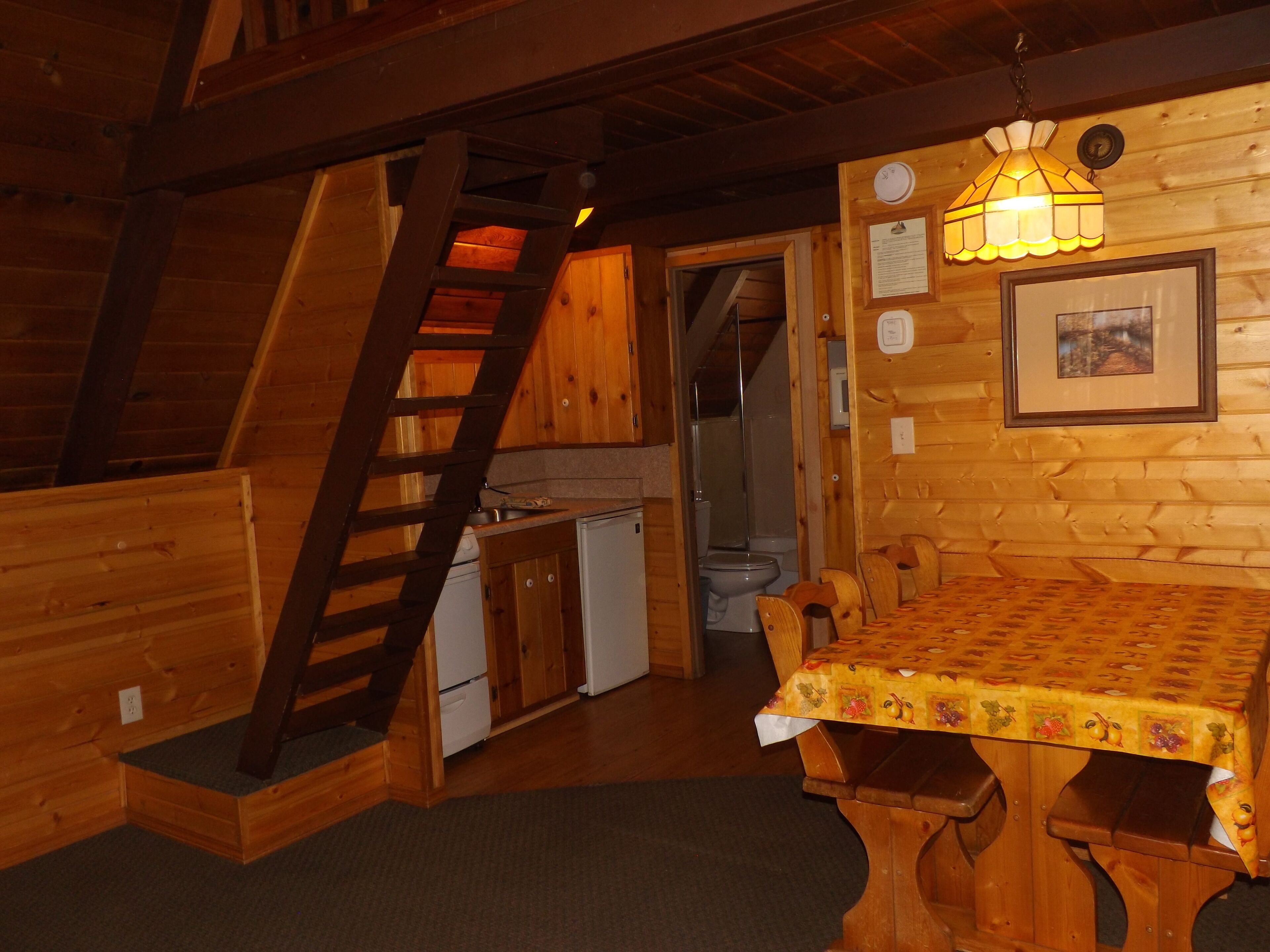 Interior de una cabaña de madera con una mesa de comedor, sillas, una zona de cocina y una escalera.
