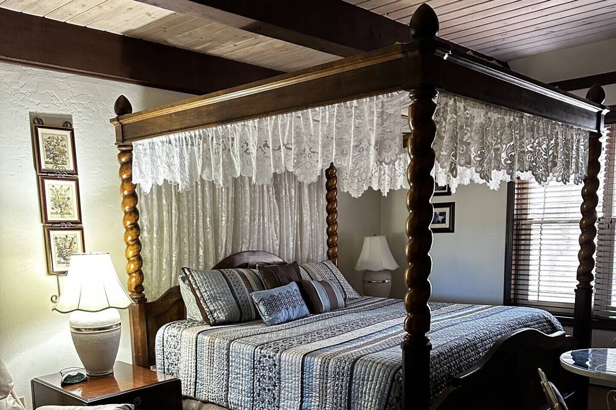 A four-poster bed with a lace canopy, a bedside table with a lamp, a chair, and a small table with a lamp.