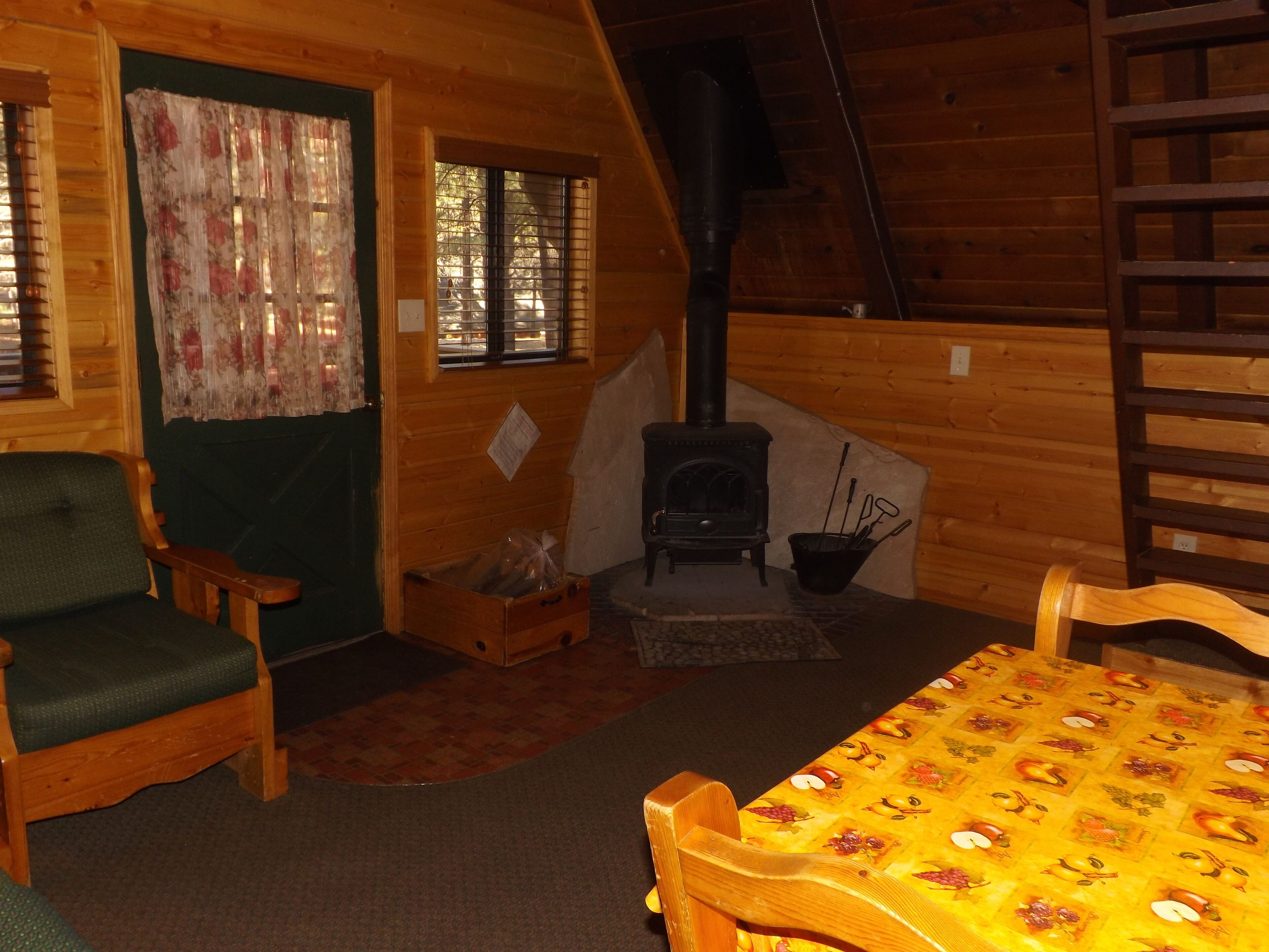 A cozy cabin interior with a wooden interior, a fireplace, a dining table with a fruit pattern tablecloth, and a green upholstered chair.