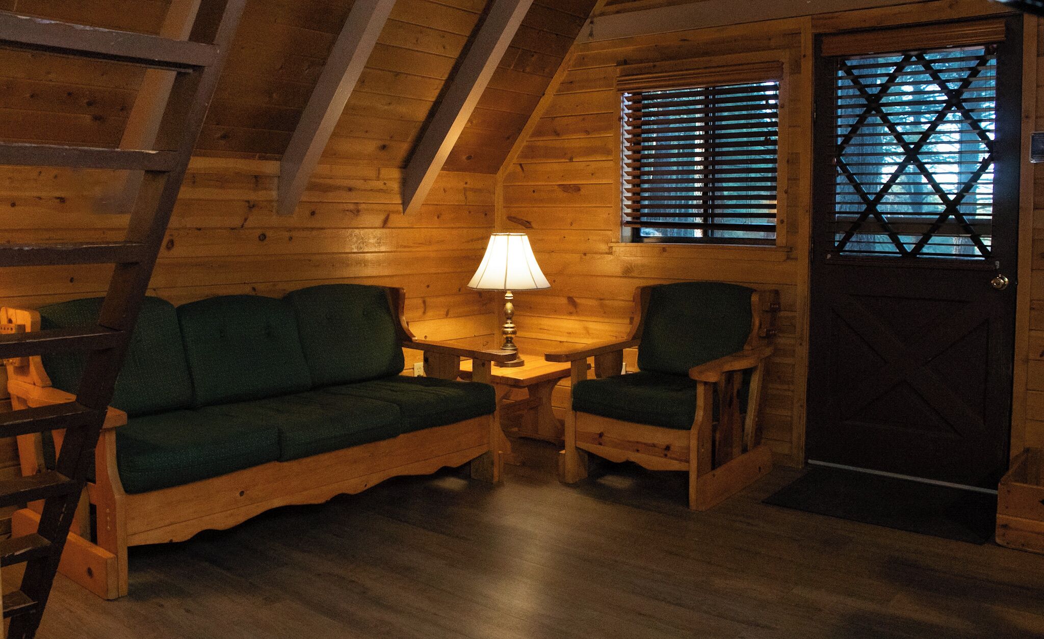 Ein gemütliches Blockhaus-Interieur mit Holzbalkendecke, einem grünen Sofa, zwei Stühlen, einem kleinen Tisch mit Lampe und einem Fenster mit Jalousien.