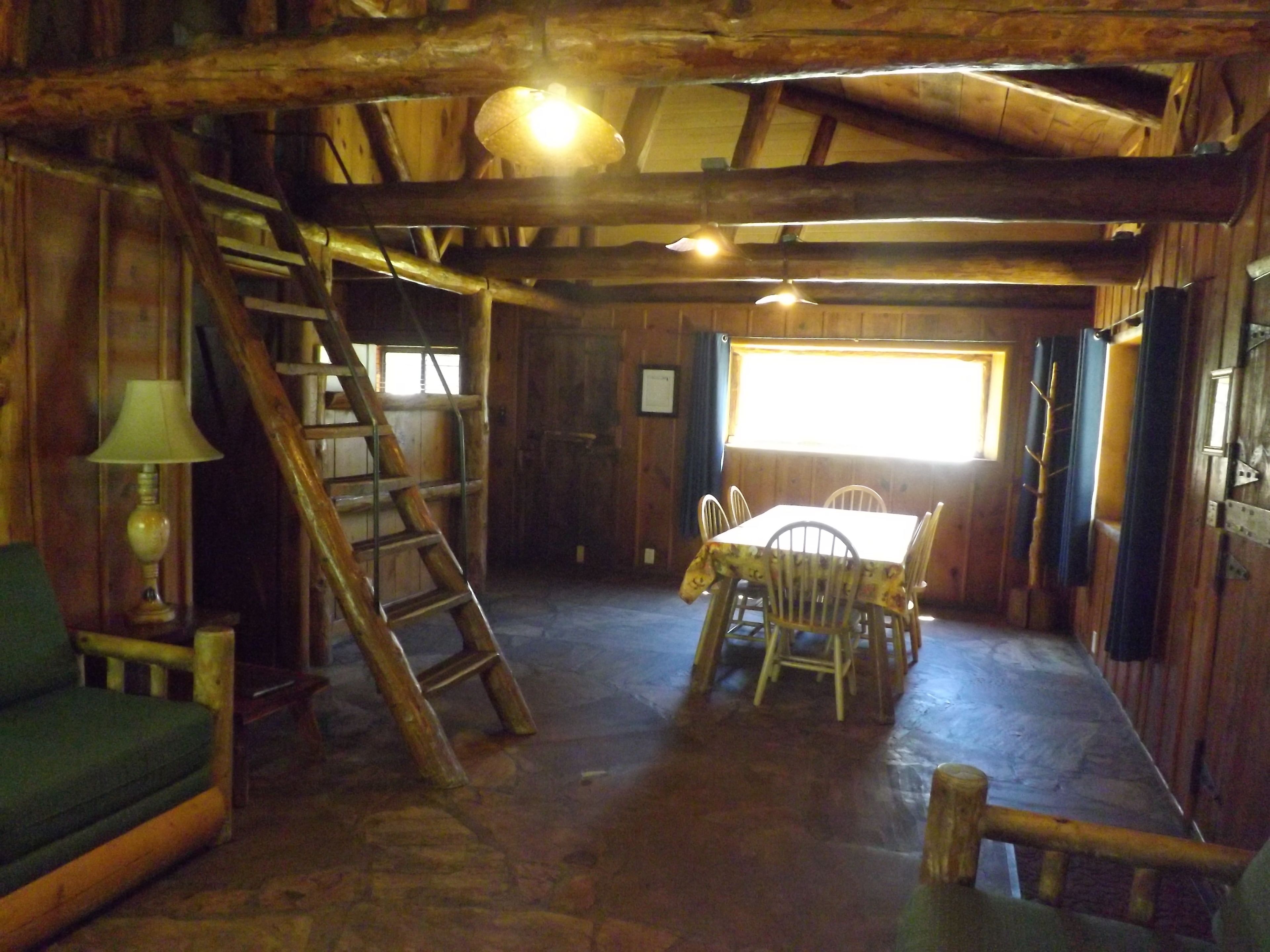 Un interior rústico de madera con una mesa de comedor, sillas, una escalera, un sofá y un ventanal que deja entrar luz natural.
